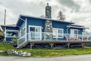 Ein zweistöckiges Haus mit einem Steinkamin, einer Terrasse mit Stühlen und einem wolkenlosen Himmel.