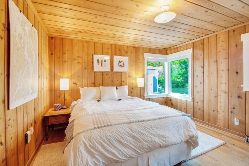 Een slaapkamer met houten lambrisering, een bed, nachtlampjes en een raam met uitzicht op groen.