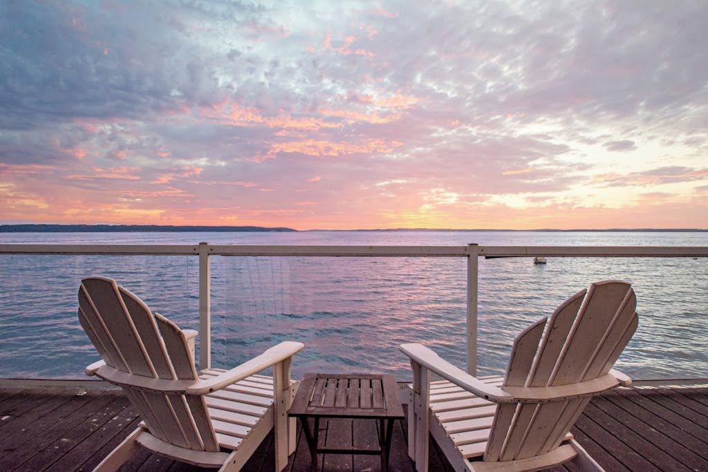 Two Adirondack chairs facing a wooden deck with a view of a body of water and a sunset.