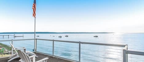 Una terraza de madera con sillas blancas y una bandera estadounidense, con vistas a un agua tranquila donde se ven barcos.