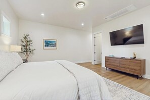 A bedroom with a bed, a television mounted on the wall, a wooden dresser, and a door leading to another room.