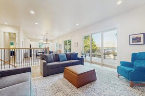 Een ruime woonkamer met een bank, poef en een blauwe fauteuil. Er is een balkon met een hekwerk en uitzicht op de omgeving.