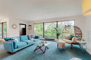 Ein modernes Wohnzimmer mit einer Couch, einem Glastisch, einem Hängesessel und Blick auf Grün hinter großen Fenstern.