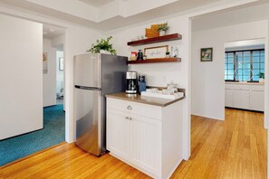 Eine moderne Küche mit einem Edelstahlkühlschrank, weißen Schränken und einer Holz Arbeitsplatte. Darüber befinden sich Regale mit Dekoration und eine Kaffeemaschine.