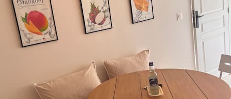 Una mesa redonda de madera con una botella y un posavasos, un banco acolchado con un plaid estampado, e ilustraciones enmarcadas de frutas en la pared.