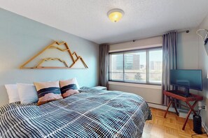 Ein Schlafzimmer mit Bett, Fernseher auf einem Schrank, einem Fenster mit Vorhängen und einer Wandleuchte.