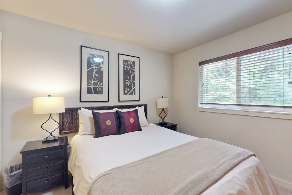 Une chambre d’hôtel avec un lit, deux tables de chevet, une fenêtre avec des stores et deux tableaux encadrés accrochés au mur.