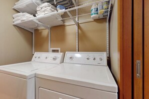 A laundry area with two white washing machines and a shelf above holding folded towels and laundry supplies. - Forest 3BR in Shoreline with patio & yard (Shoreline)