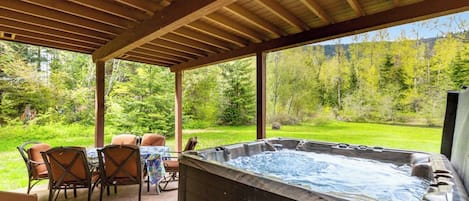 A hot tub under a wooden pergola with a table and chairs, surrounded by a lush green landscape.
