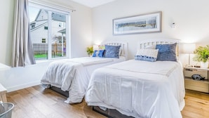 Et soveværelse med to senge, et vindue med gardiner, et billede i ramme på væggen og et natbord med en lampe.