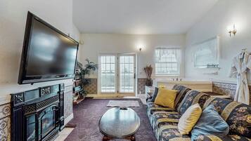 A living room with a fireplace, a flat-screen TV, a sofa with patterned cushions, and a window with blinds.