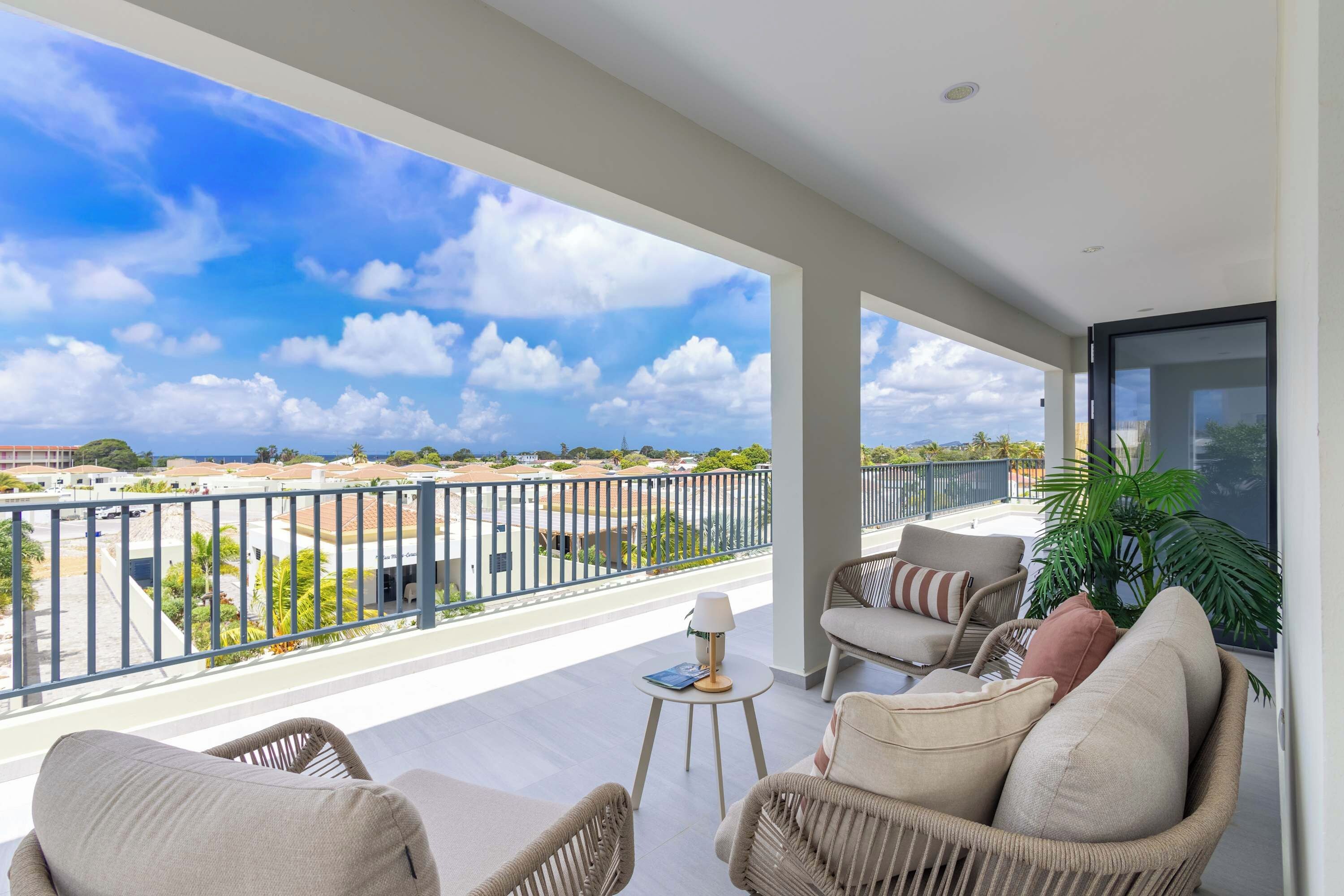 A balcony with wicker furniture, a small table, and a view of a coastal area with buildings and greenery.