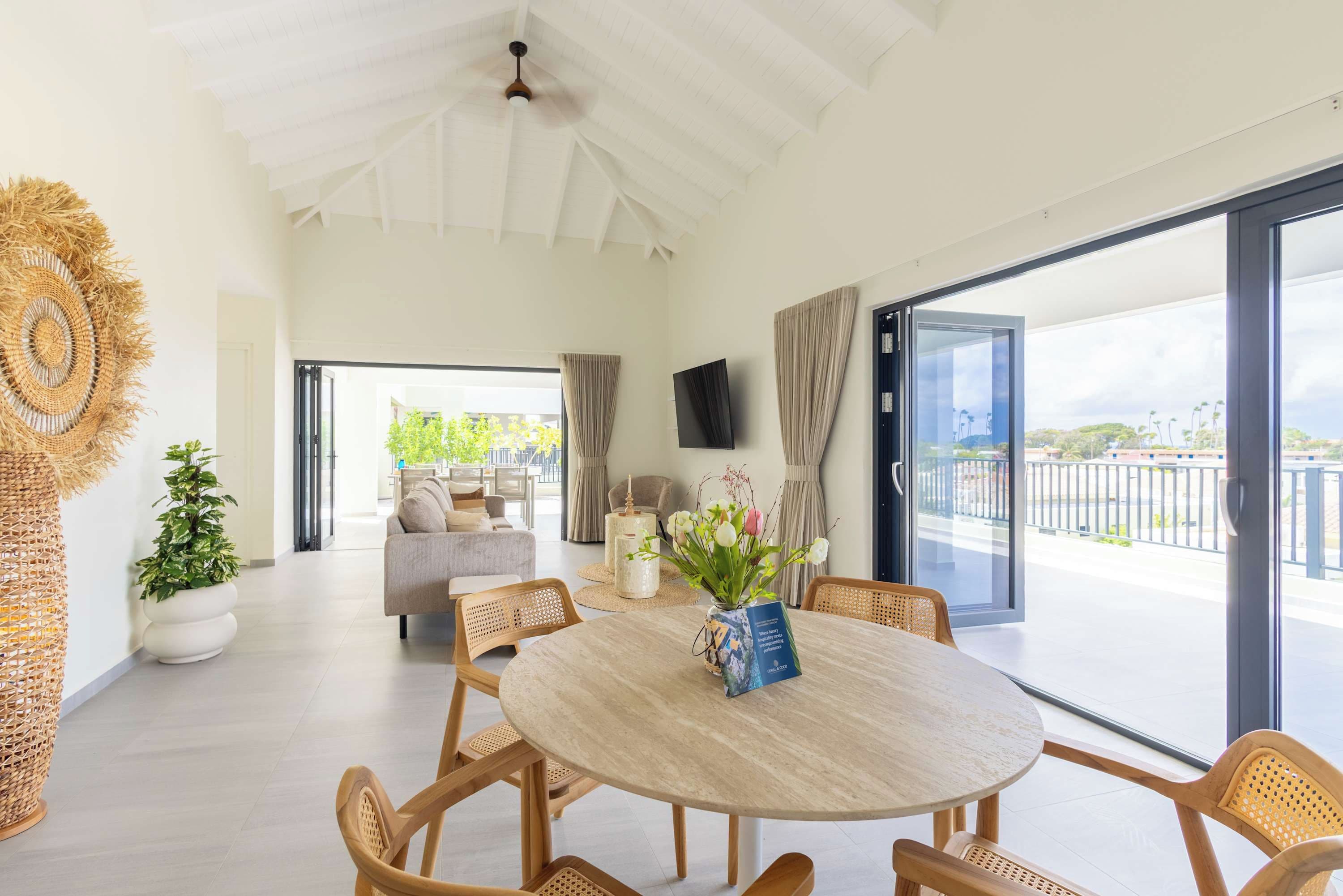 A spacious living area with a round table, wicker chairs, a sofa, and a view through large glass doors to the outside.