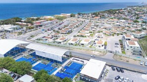 A coastal residential area with a mix of single and multi-story houses, a main road, and a large sports facility with blue courts. - "Altura" | Luxury 3BR Penthouse by Mambo Beach (Willemstad)