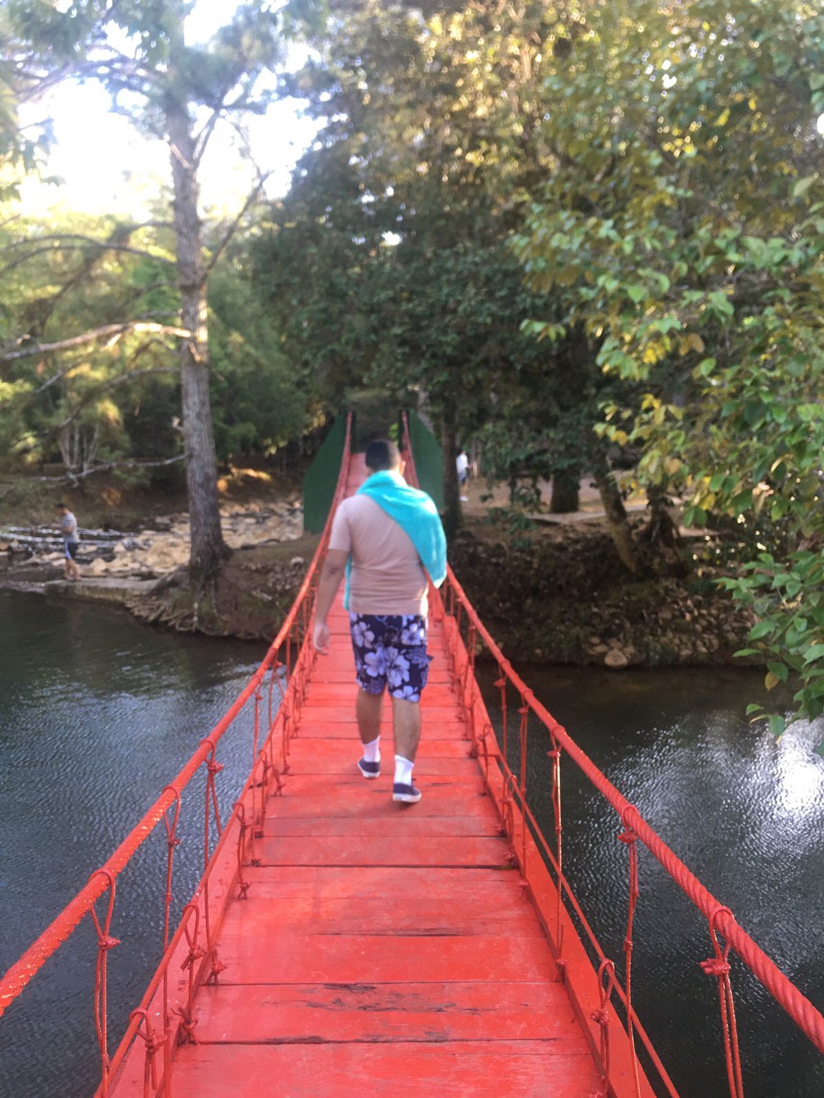 Eine Person geht über eine rote Hängebrücke, die über ein Gewässer führt, umgeben von Bäumen.