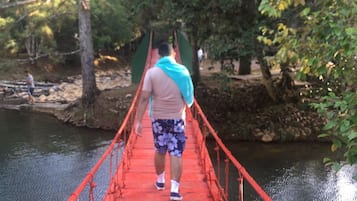Eine Person geht über eine rote Hängebrücke, die über ein Gewässer führt, umgeben von Bäumen.