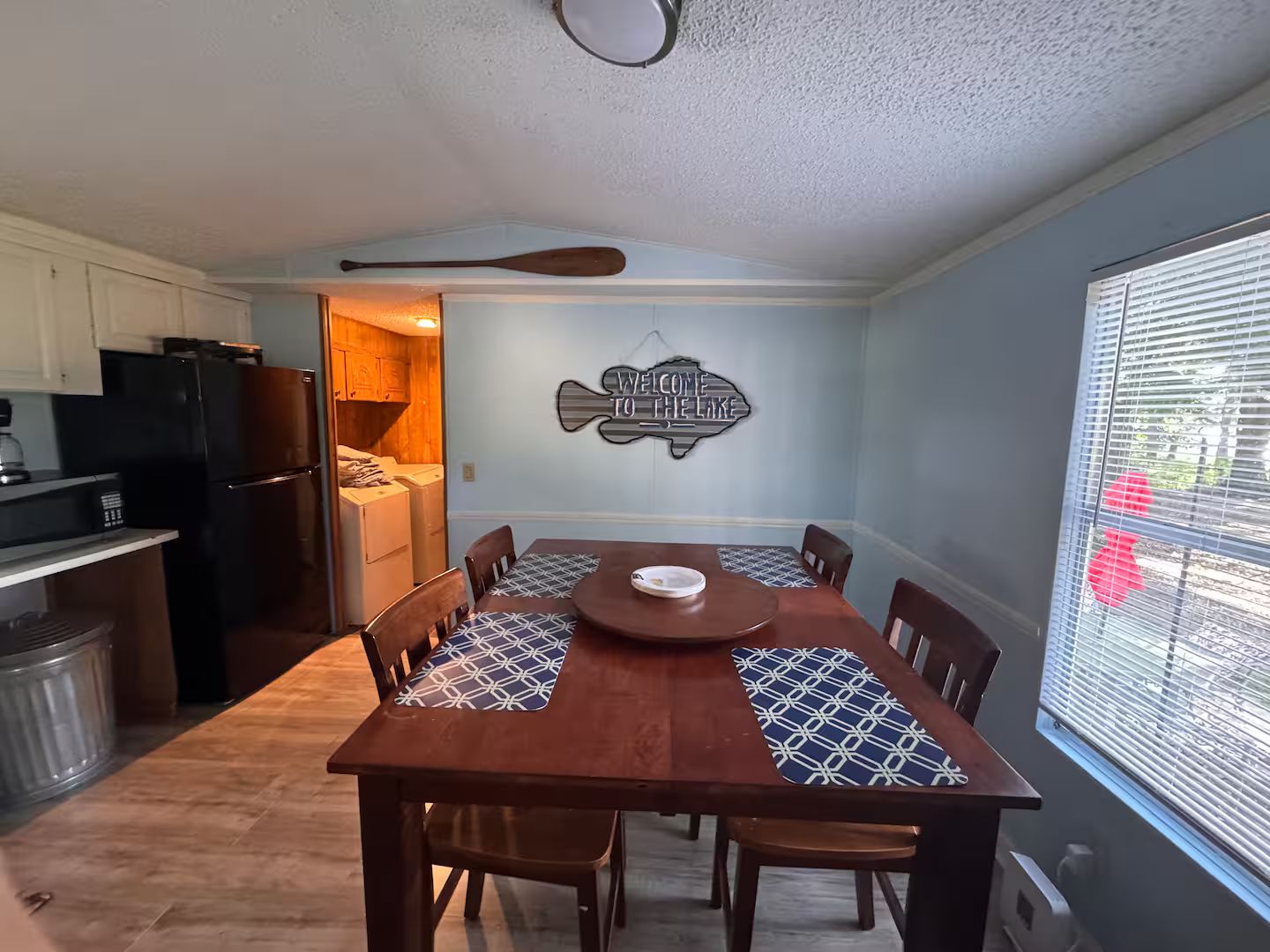 A dining area with a wooden table and chairs, a kitchen in the background, and a wall sign that reads 'WELCOME TO THE LAKE'.