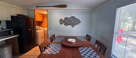A dining area with a wooden table and chairs, a kitchen in the background, and a wall sign that reads 'WELCOME TO THE LAKE'.