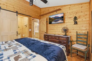 A wooden cabin room with a bed, dresser, chair, and a painting on the wall. - Bear Essentials & Hot Tub (Blairsville)