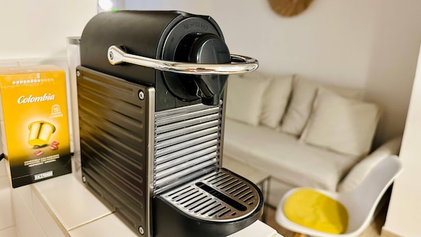 Une machine à café dotée d’un bac de récupération, un compartiment pour les capsules de café étiqueté « Colombie », et une chaise blanche avec un coussin jaune.