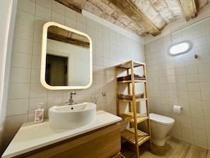 Ein Badezimmer mit einem weißen Waschbecken, einer Holzablage, einem Spiegel und einer Toilette.