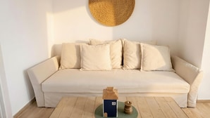 Un canapé aux couleurs neutres, avec une tapisserie murale tissée au-dessus et un petit objet en forme de maison posé sur le sol.