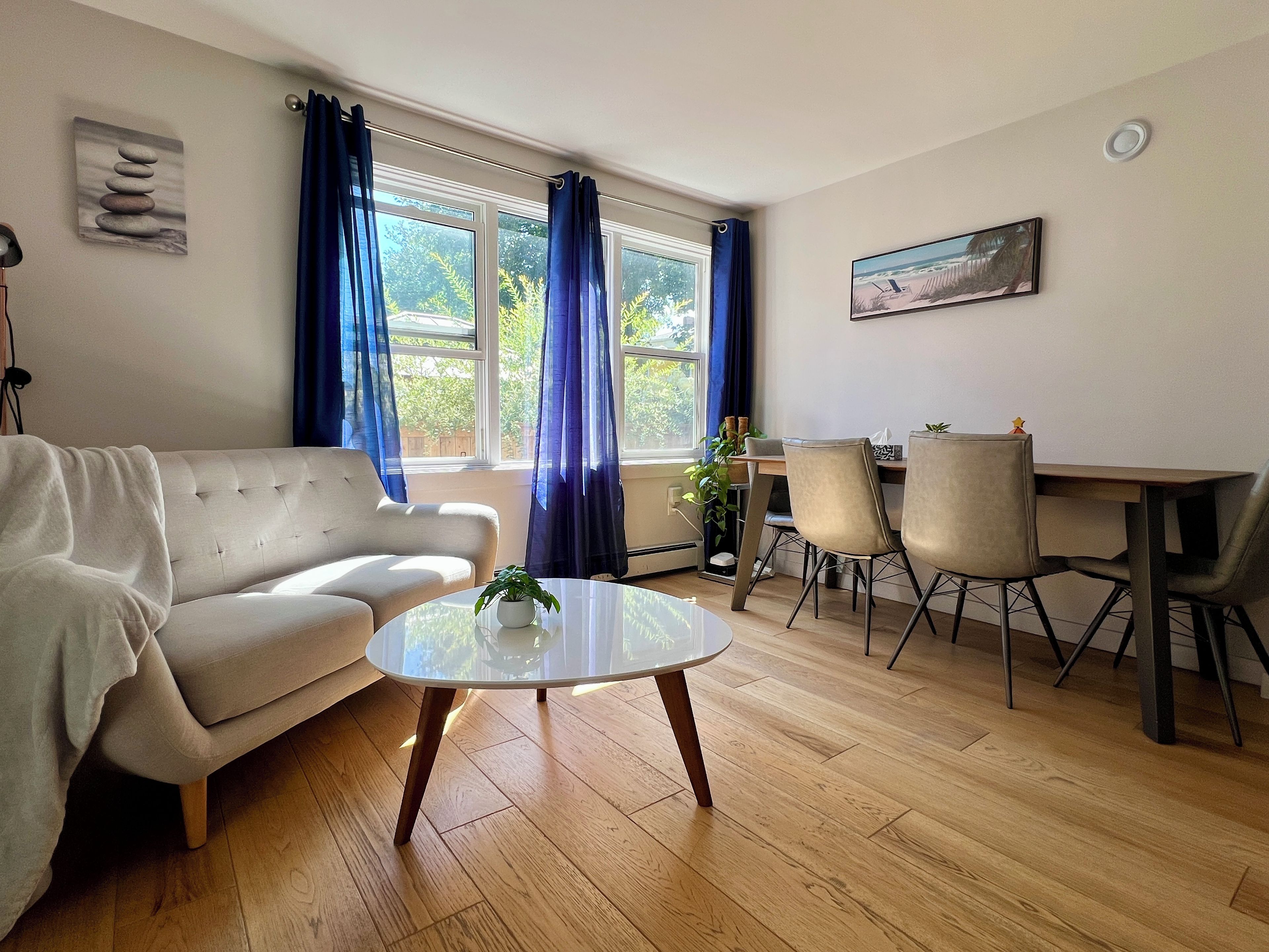 Uma sala de estar com sofá, mesa de jantar com cadeiras, mesa de centro e um quadro na parede.