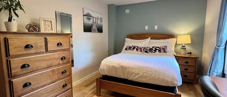 Un dormitorio con cama de madera, un aparador, una mesita de noche con lámpara, una planta en maceta y un cuadro en la pared.