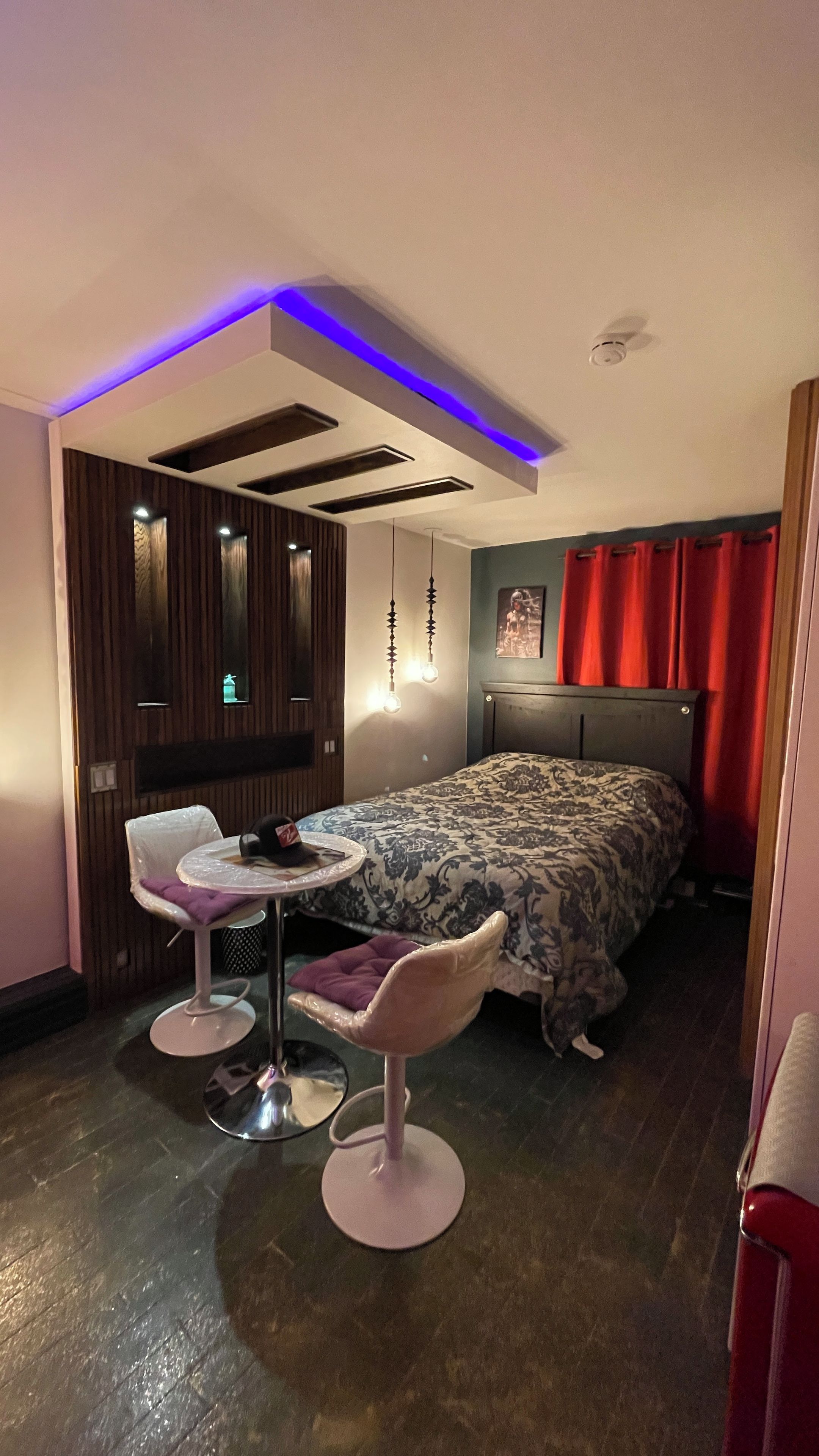 Un dormitorio moderno con una cama, una mesita redonda pequeña, dos sillas, un aplique de techo y un televisor instalado en la pared.
