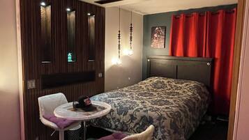 Un dormitorio moderno con una cama, una mesita redonda pequeña, dos sillas, un aplique de techo y un televisor instalado en la pared.