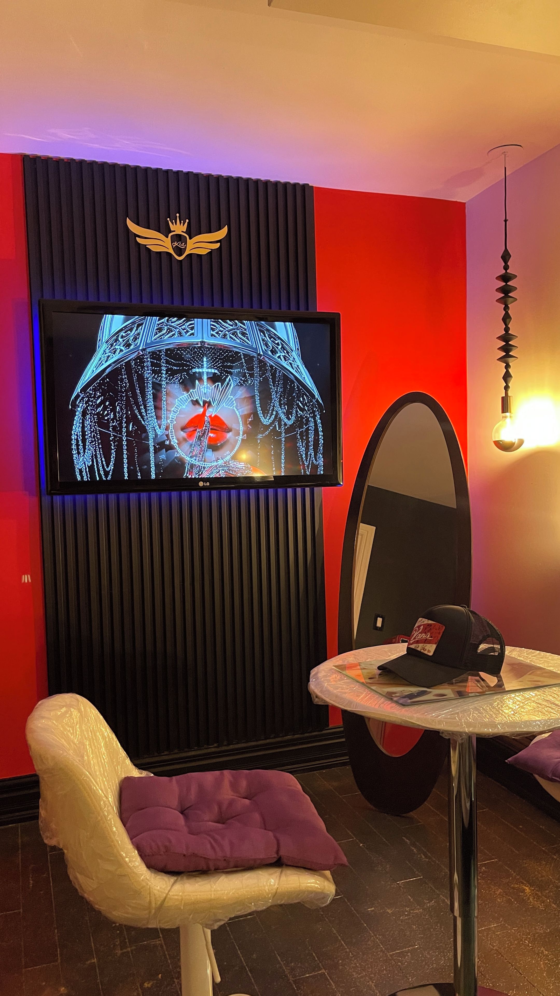 Una habitación con una televisión montada en un panel negro, un espejo redondo, una mesita con un sombrero y un sillón acolchado.