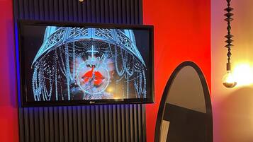 Una habitación con una televisión montada en un panel negro, un espejo redondo, una mesita con un sombrero y un sillón acolchado.