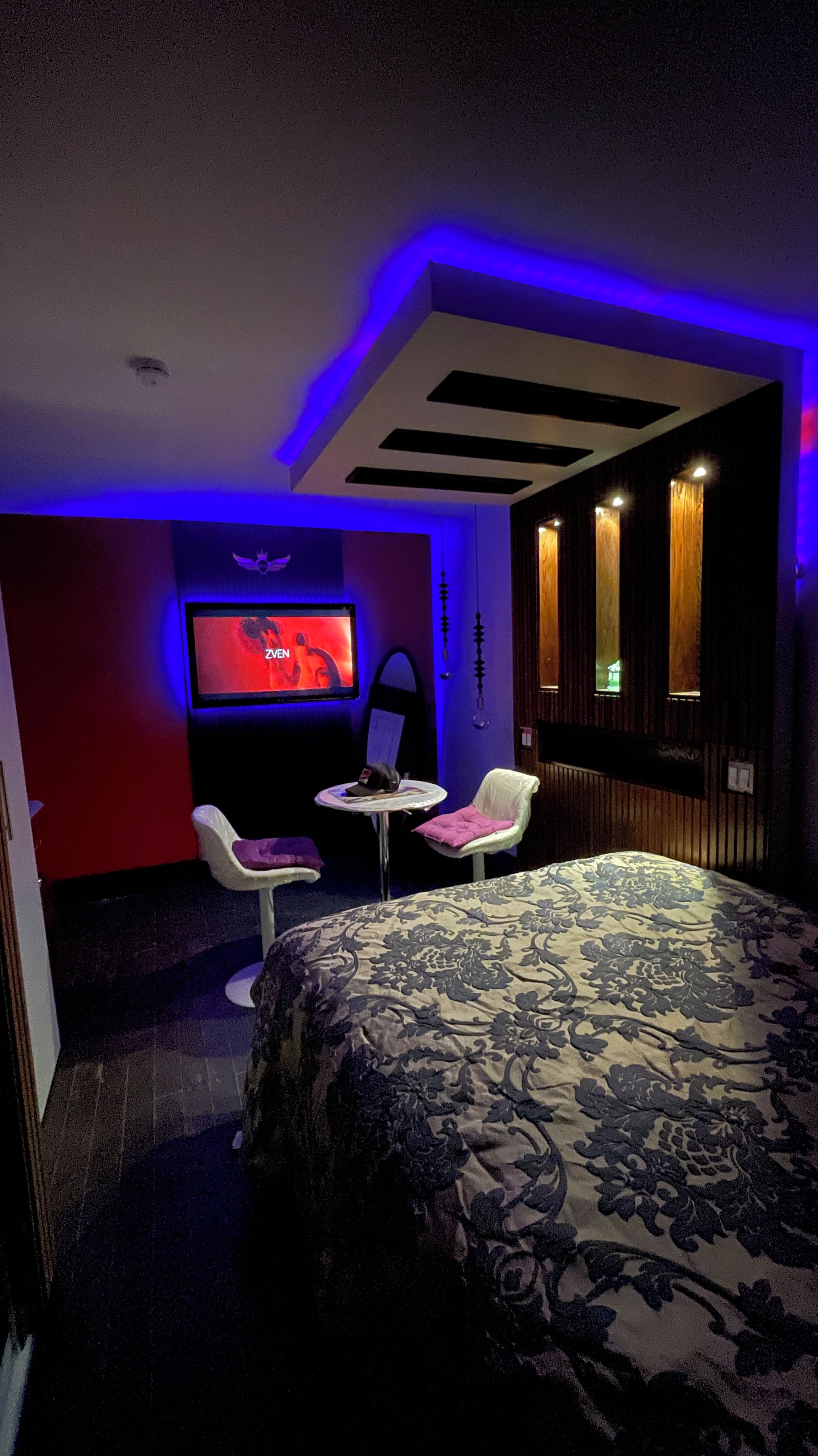 Une chambre d’hôtel comprenant un lit, deux chaises, une petite table, un téléviseur et une étagère fixée au mur.
