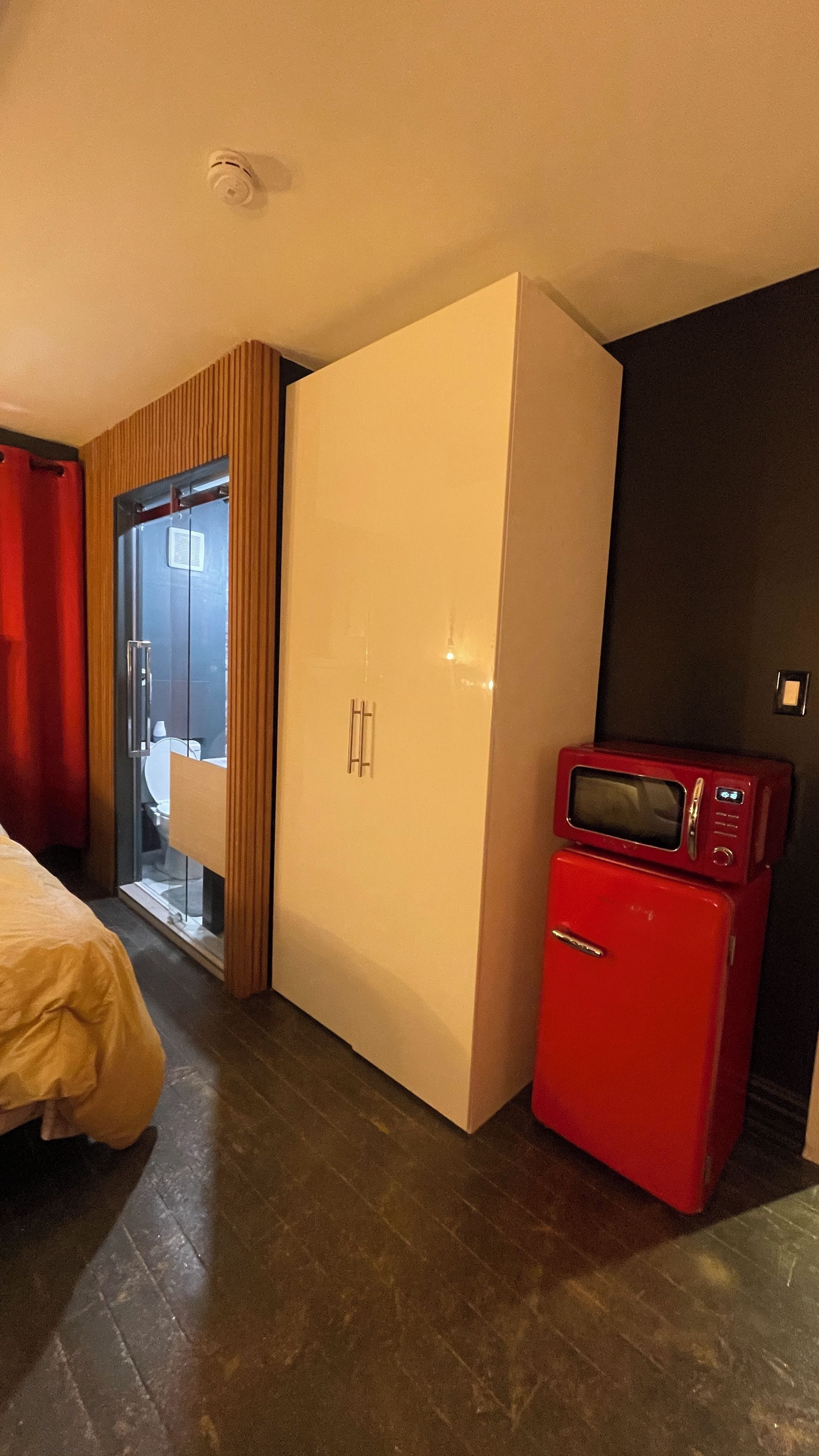 Une chambre d’hôtel avec un lit, une armoire, un four à micro-ondes rouge et une salle de bain visible à travers une porte en verre.