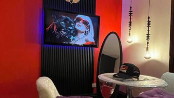 Ein modernes Zimmer mit einem an einer schwarzen Platte befestigten Fernseher, einem runden Spiegel und einem Tisch mit einem violetten Kissen.
