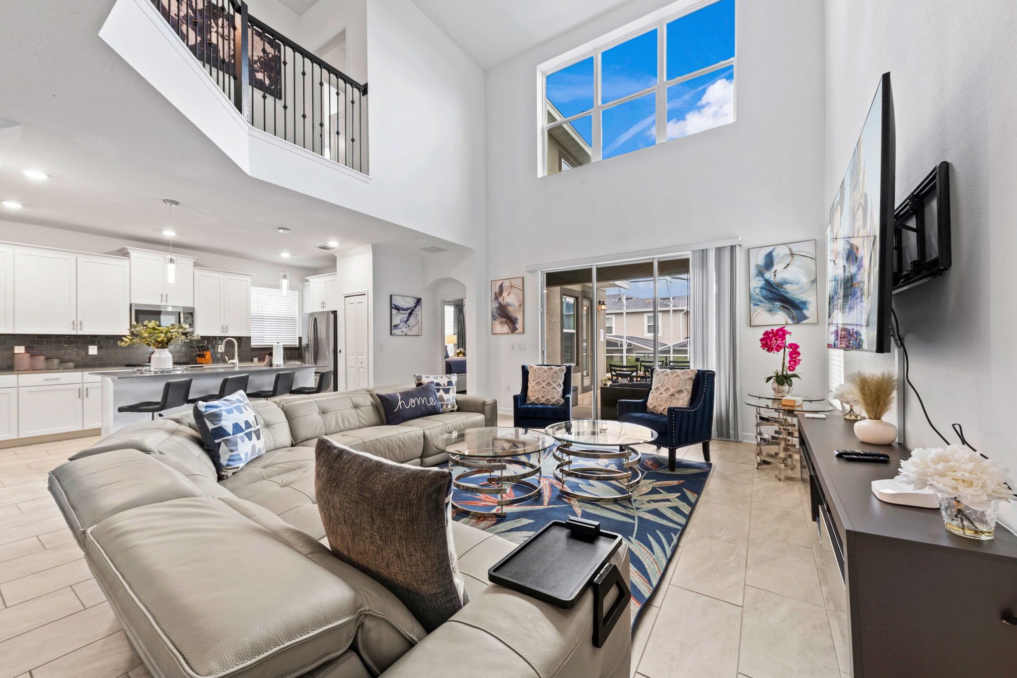 A spacious living area with a high ceiling, a kitchen in the background, and a large window providing natural light.