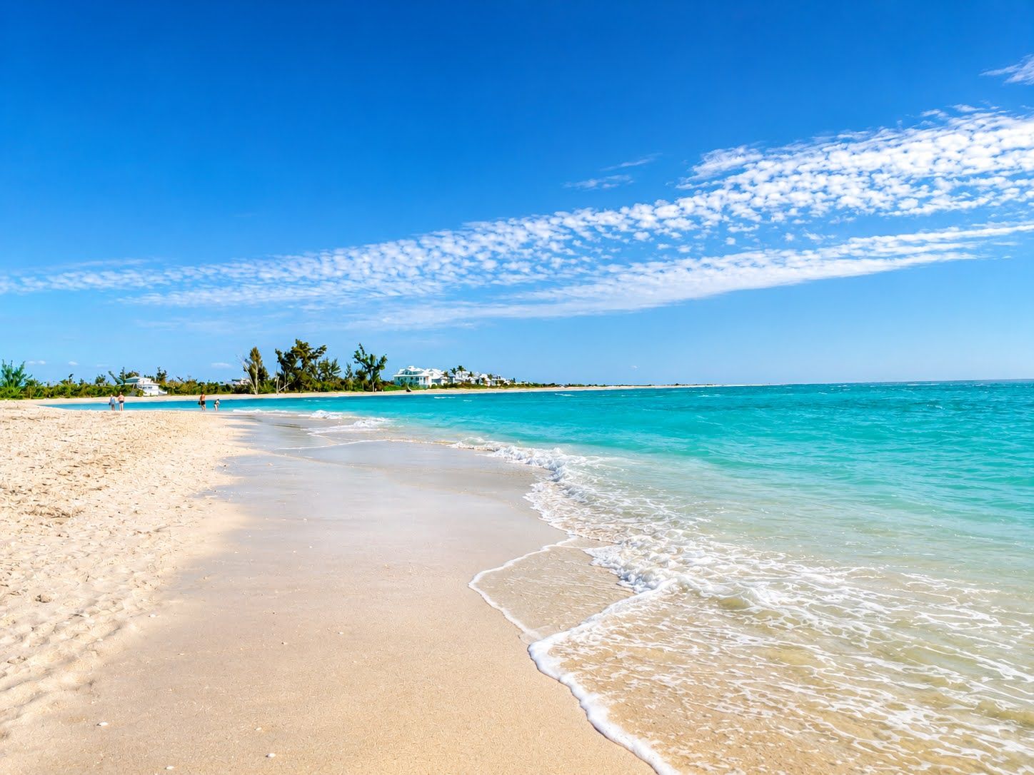 Een strand met helder turquoise water, een zandstrand en enkele gebouwen in de verte.