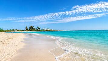 Een strand met helder turquoise water, een zandstrand en enkele gebouwen in de verte.