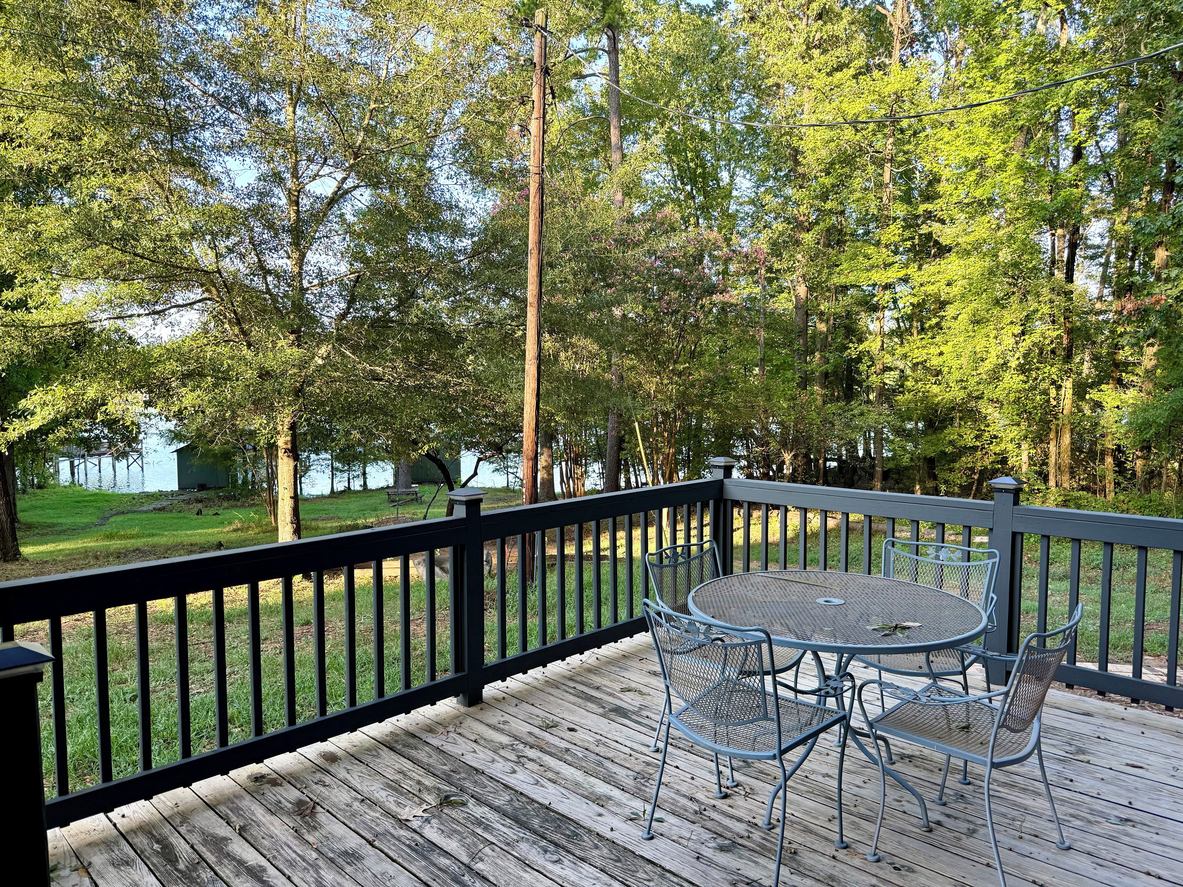 A wooden deck with metal chairs and a table, overlooking a grassy area with trees and a body of water.