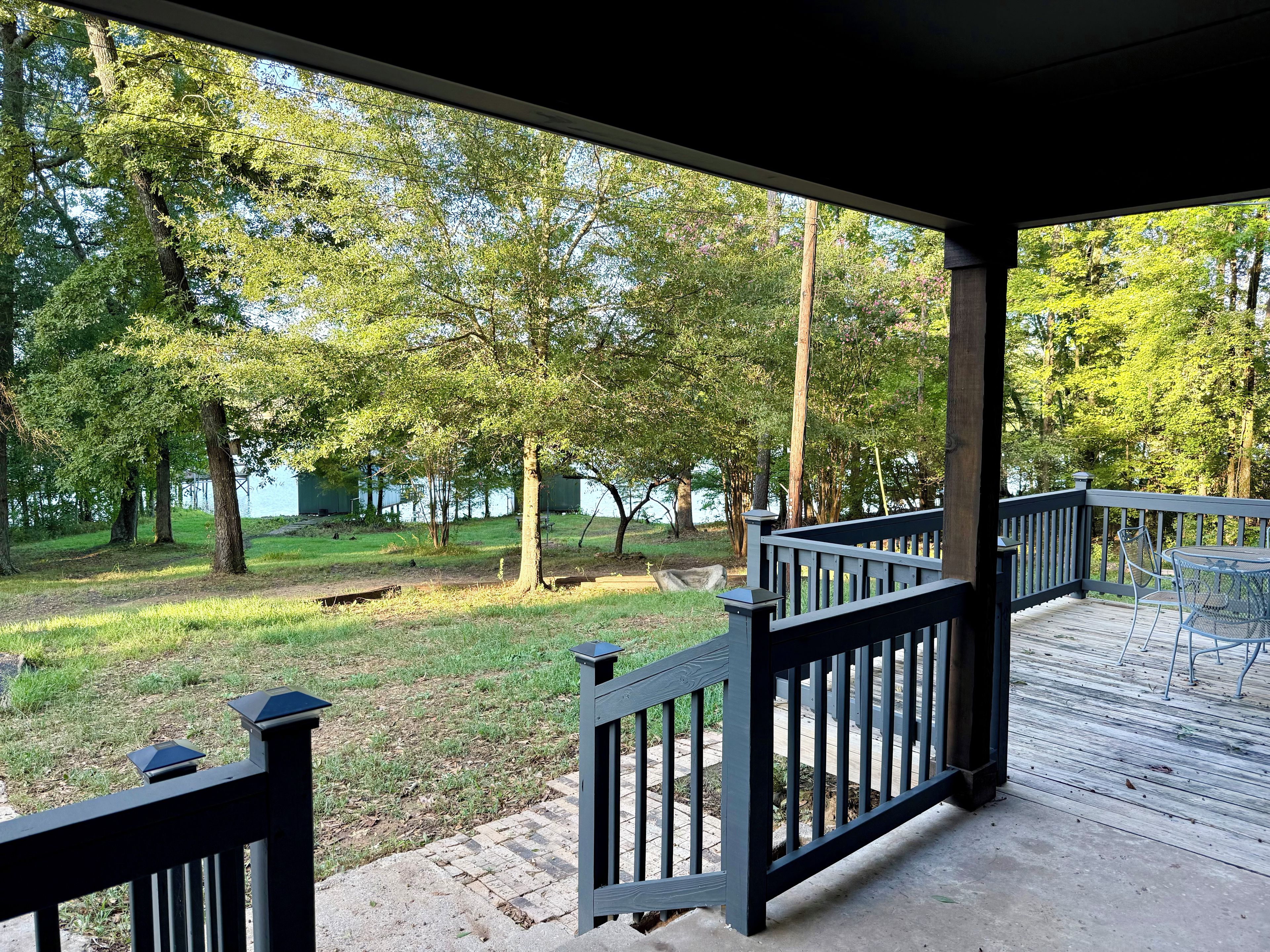 A covered outdoor deck with a railing, overlooking a grassy area with trees and a body of water in the background.