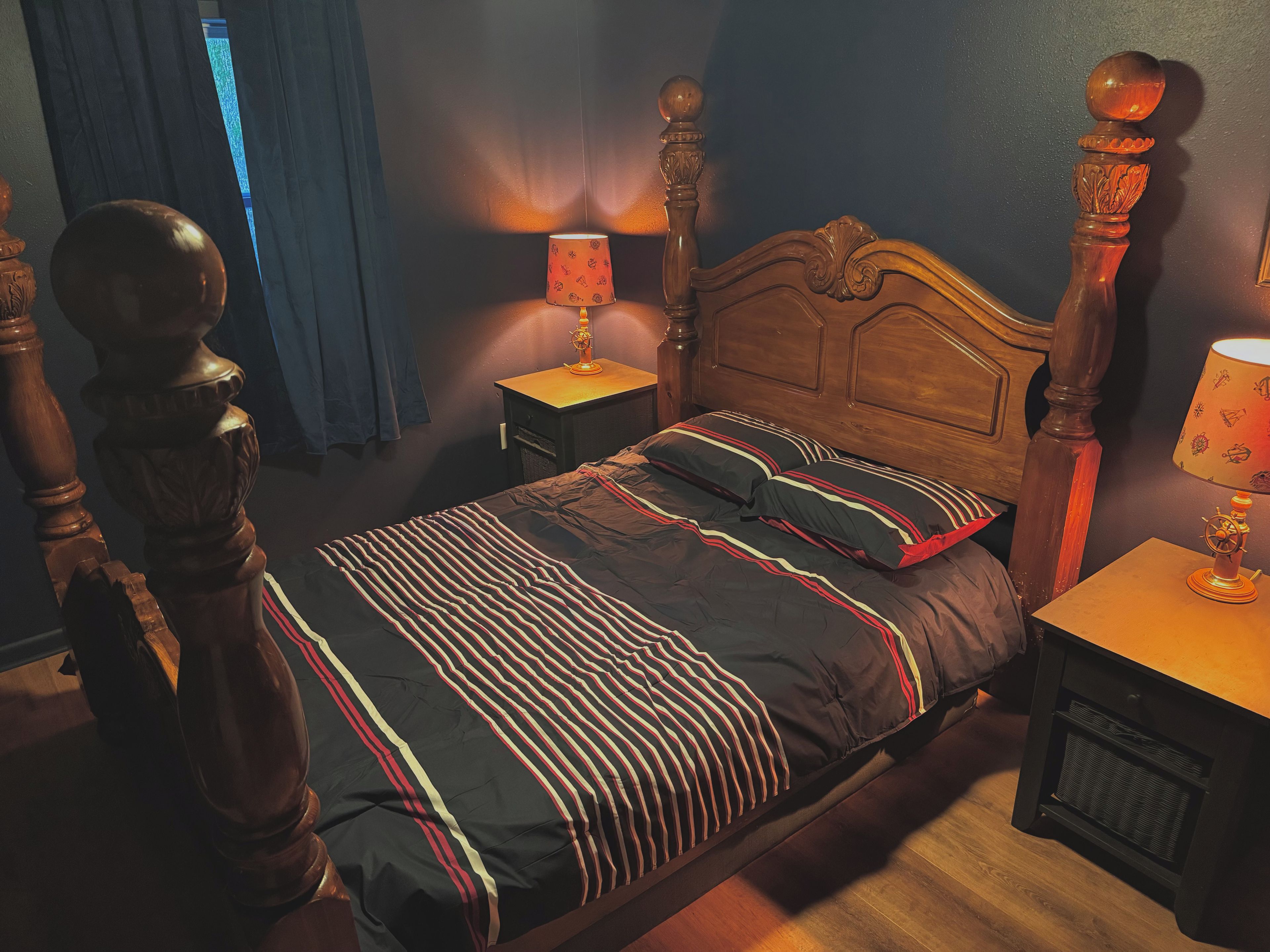 A bedroom with a wooden bed, two bedside tables with lamps, a window with curtains, and a wooden floor.