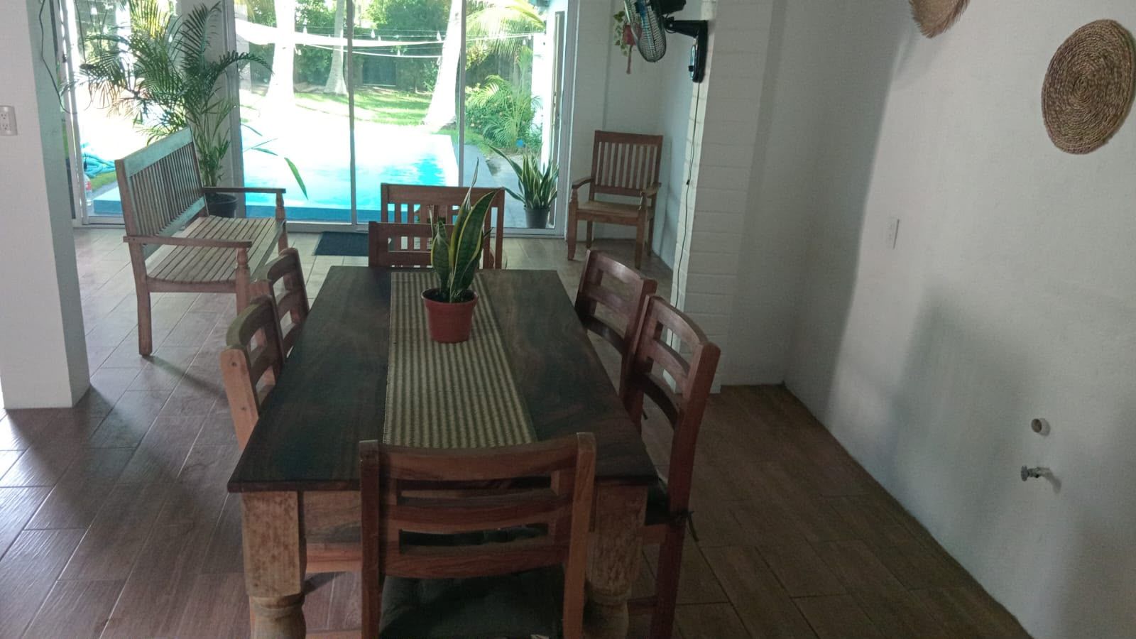 Spiseområde med et træbord og stole, en potteplante og udsigt til en swimmingpool gennem en glasdør.