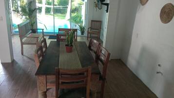 Spiseområde med et træbord og stole, en potteplante og udsigt til en swimmingpool gennem en glasdør.