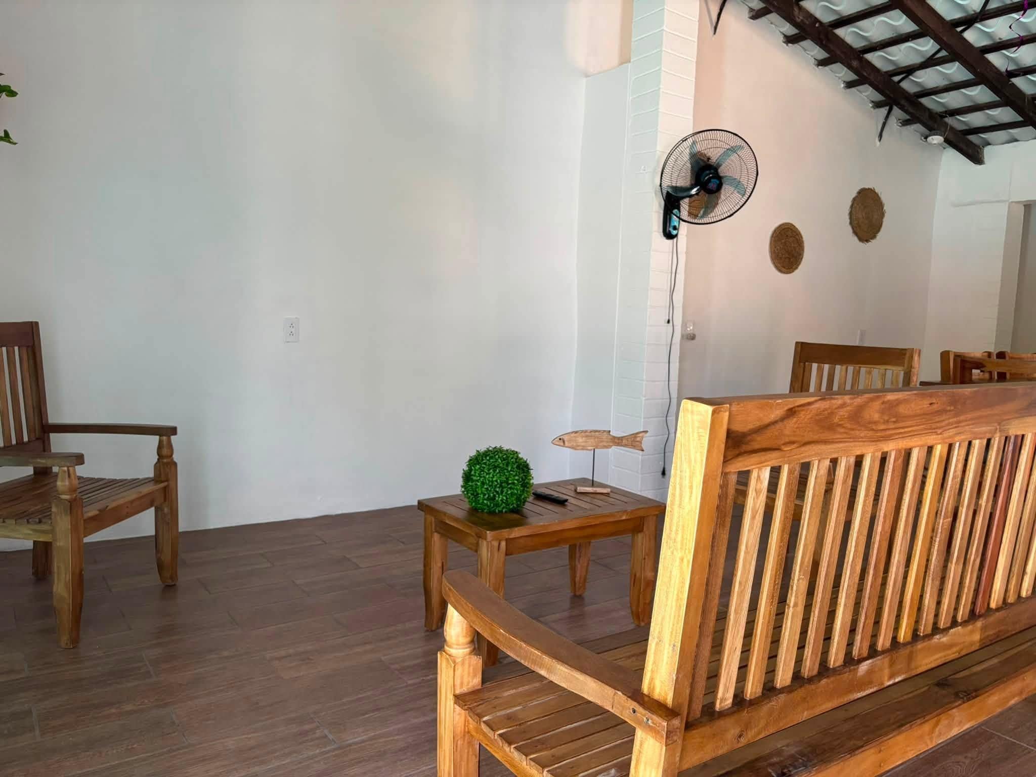 Quarto com móveis de madeira, incluindo um sofá, cadeiras, uma mesa pequena com um objeto decorativo e um ventilador fixado na parede.