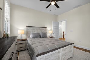 A bedroom with a bed, bedside tables, a ceiling fan, and a door leading to another room. - Bayside Wayside - 3BR Home, Walk to Beach + Pool (Cape Charles)