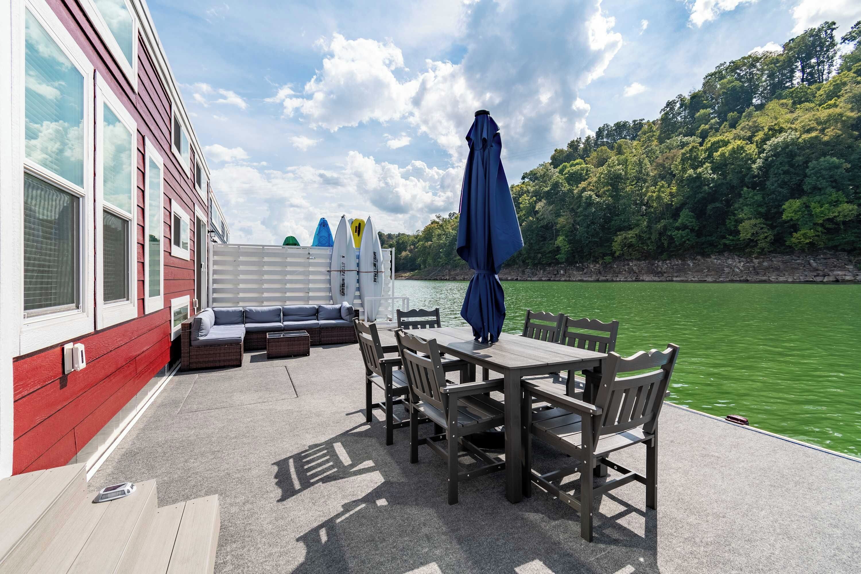 Eine Terrasse mit Gartenmöbeln, Tisch und Stühlen mit Blick auf ein Gewässer, im Hintergrund Bäume.