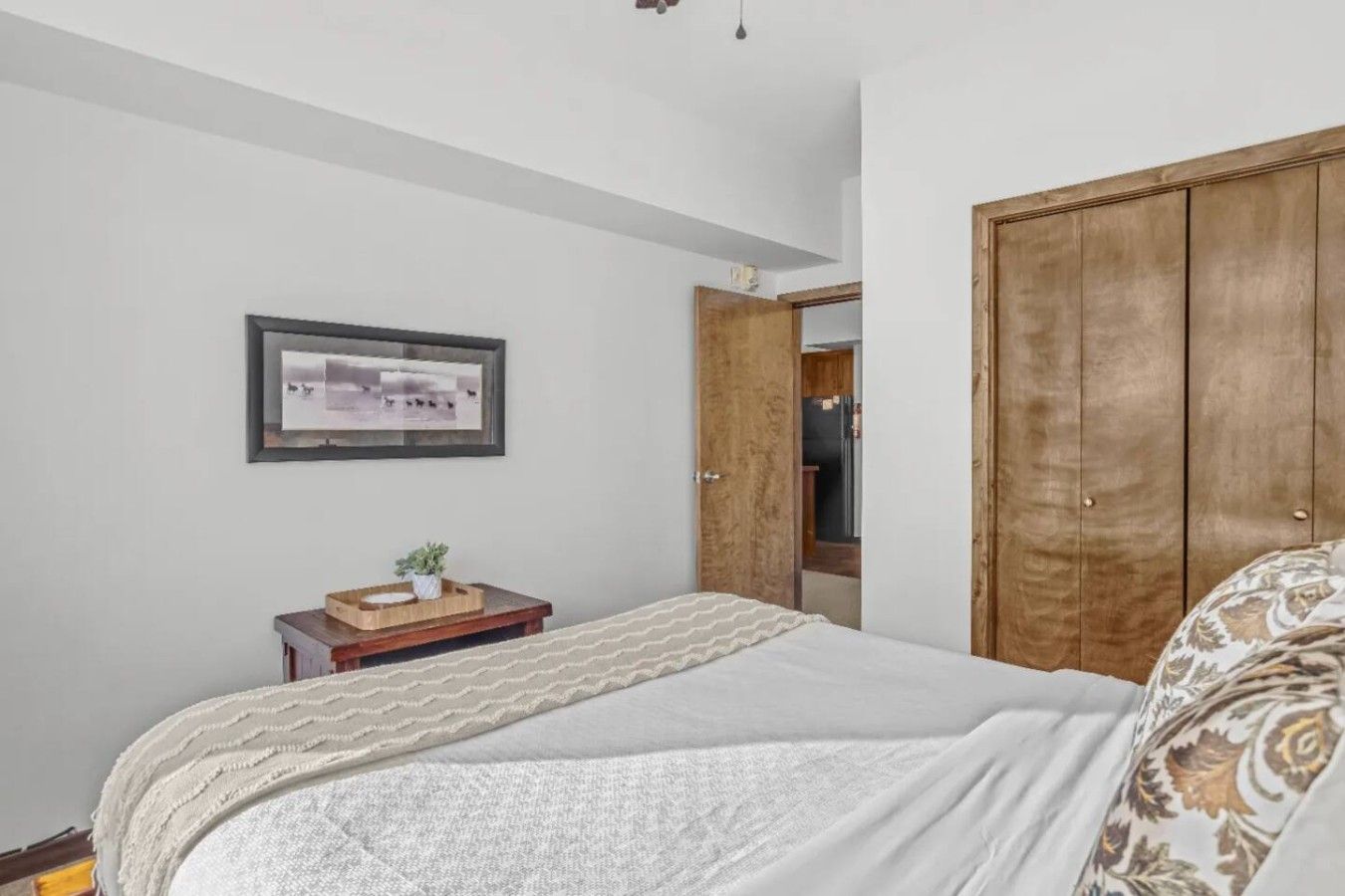 Un dormitorio con cama, una mesita de noche con una planta, un cuadro enmarcado en la pared y un armario de madera.