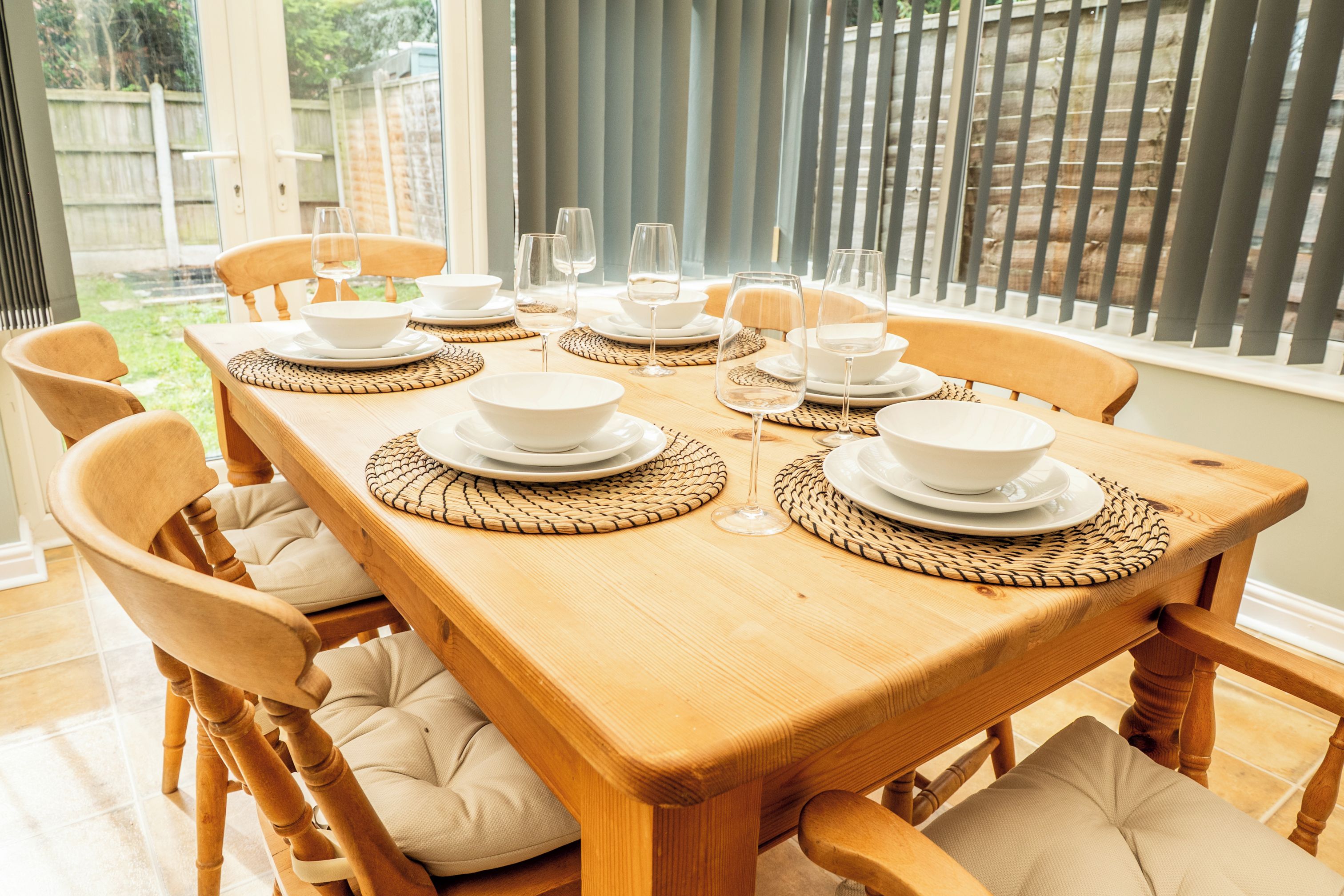 Une table à manger dressée avec de l’assiette blanche, des bols et des verres à vin, entourée de chaises en bois dotées d’assises rembourrées.