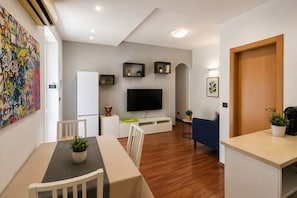 Ein modernes Wohnzimmer mit Flachbildfernseher, einem Essbereich mit Tisch und Stühlen sowie einer Küchenzeile mit Kaffeemaschine.