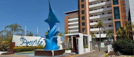 Entrada de hotel con una gran escultura azul, un letrero que dice 'Pacifika' y un edificio de varios pisos con balcones.